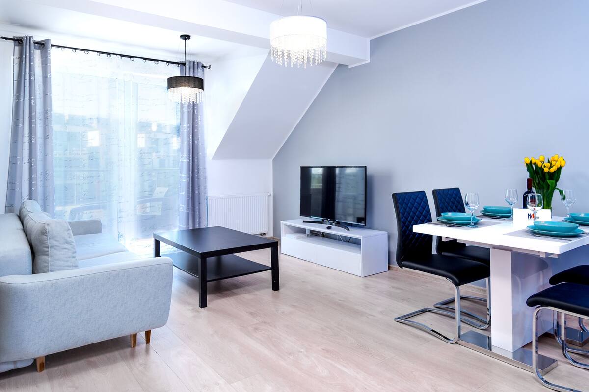 Un salon moderne avec un canapé, une table à manger dressée avec de l’assiette et des verres bleus, un téléviseur et un lustre.