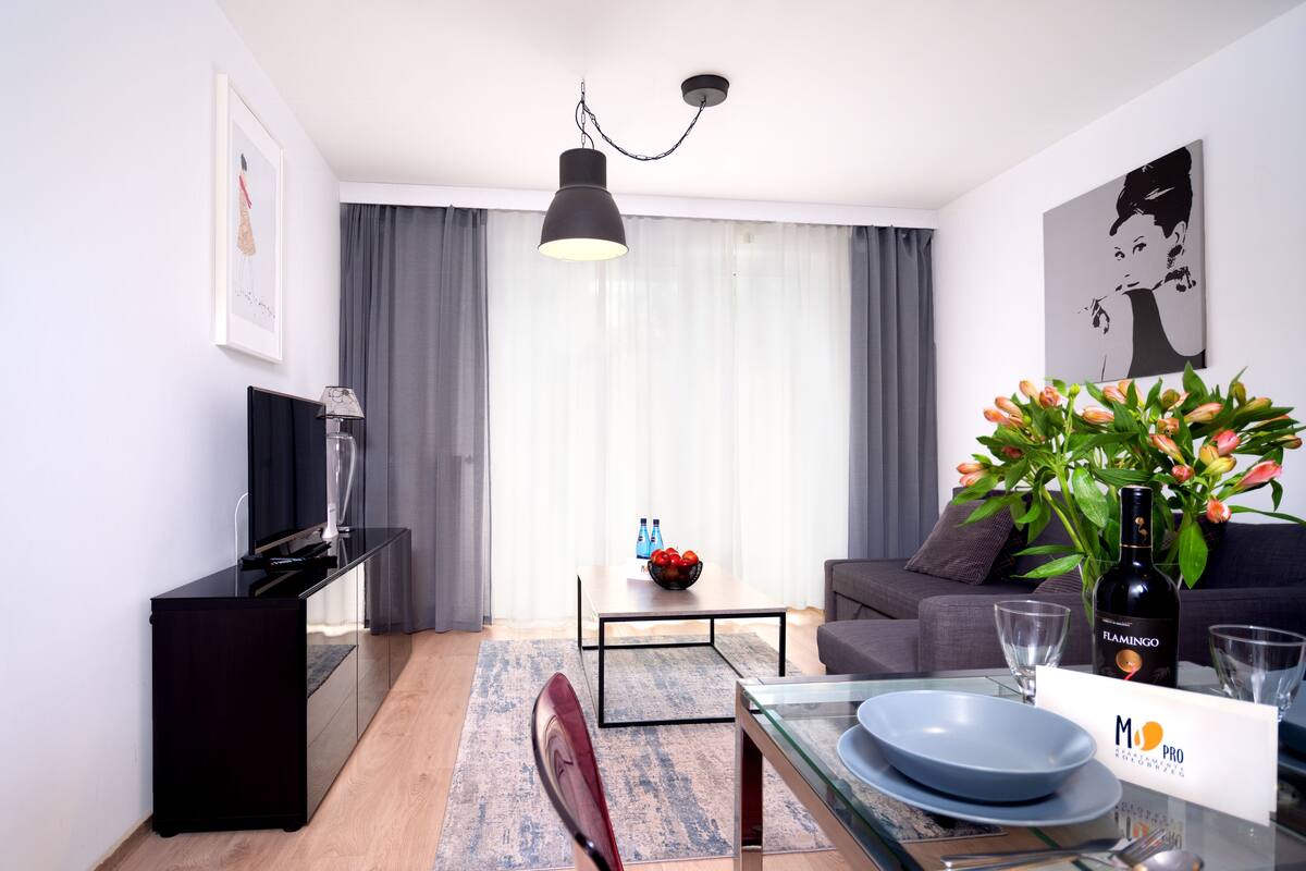 Un salon moderne avec un coin repas, comprenant un téléviseur, un canapé, une table basse avec un saladier de fruits et un vase de fleurs.