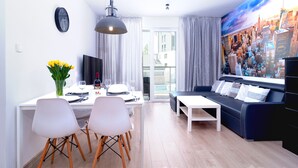 Un salon moderne avec un canapé noir, des chaises de salle à manger blanches, une table à manger dressée pour deux personnes, un téléviseur et une vue sur le paysage urbain à travers de grandes fenêtres.