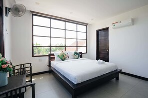 A bedroom with a large window, a bed with white linens, a bedside table with a fan, and a potted plant. - Hoi An Lotus Aroma Villa (Da Nang)