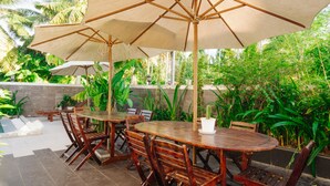 Un patio al aire libre con mesas y sillas de madera, sombrillas grandes y abundante vegetación.