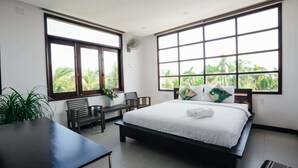 Un dormitorio con una cama grande, un escritorio y dos sillas. Amplias ventanas con vistas a palmeras.