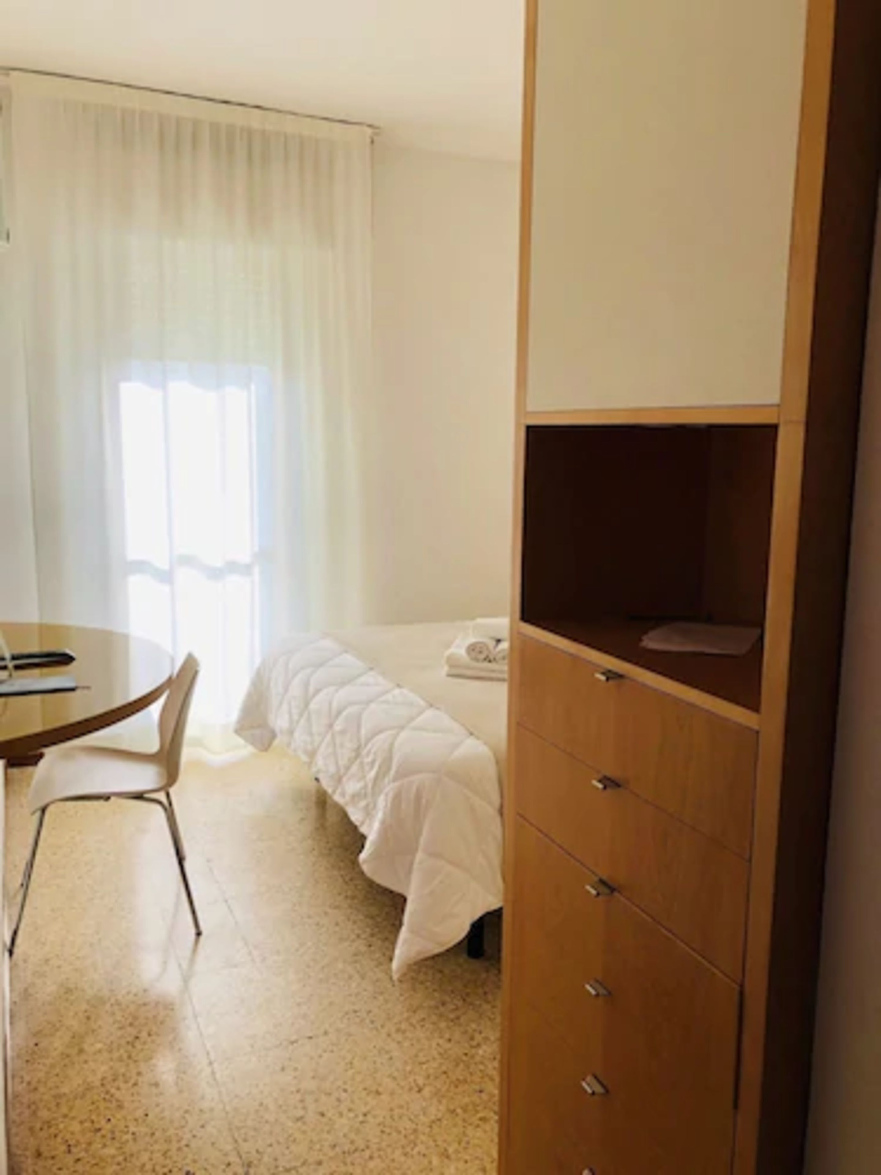 A hotel room with a bed, a wooden wardrobe, a round table with a chair, and a window with curtains.