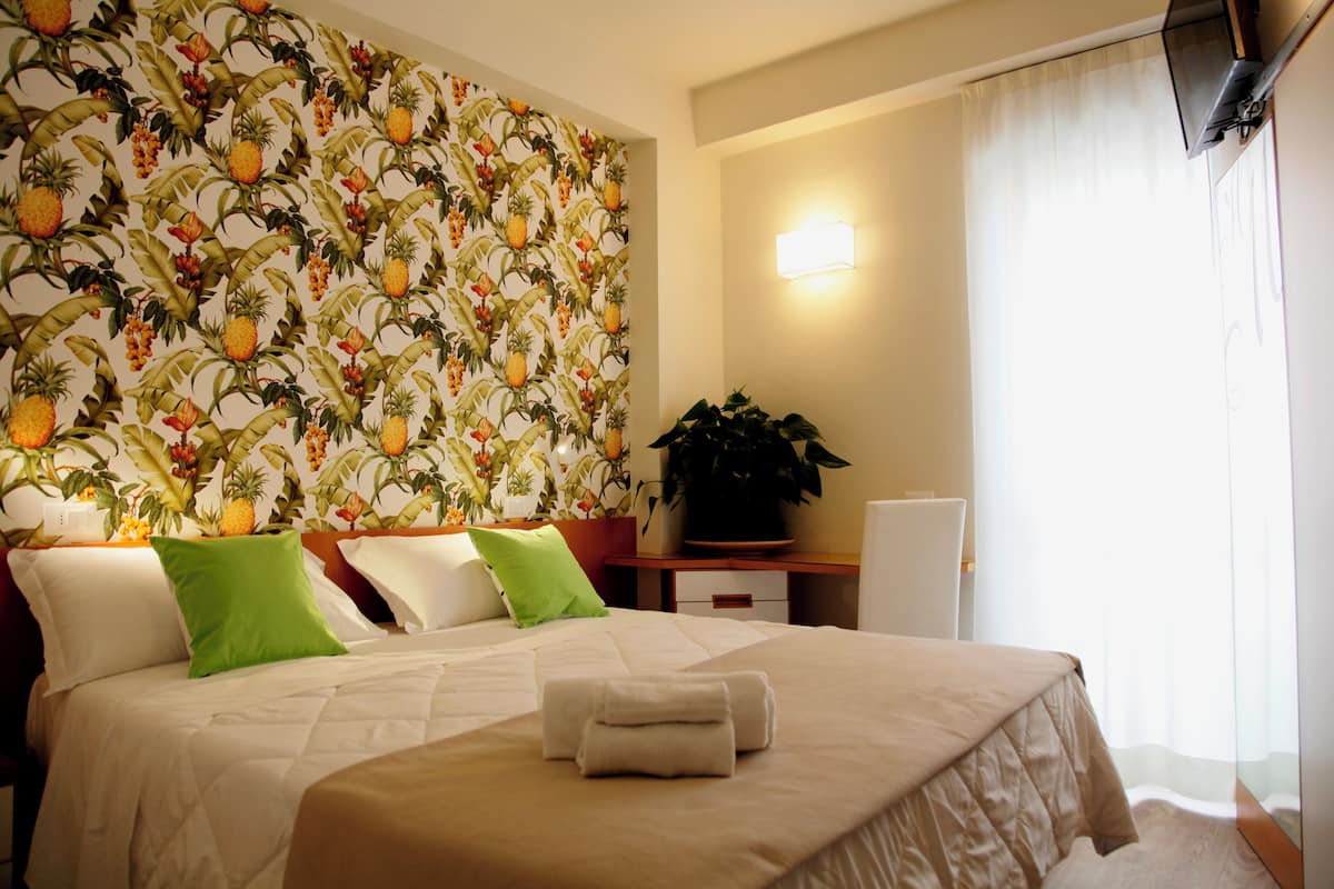 Habitación de hotel con cama, almohadas, mesita de noche, una planta y una pared con diseño de frutas.