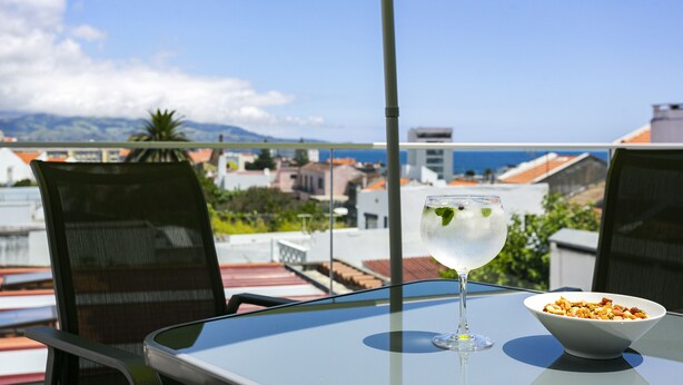 A table with a glass of water and a bowl of nuts, overlooking a cityscape with buildings and mountains. - Home Azores – Casas da Ladeira (Ponta Delgada)