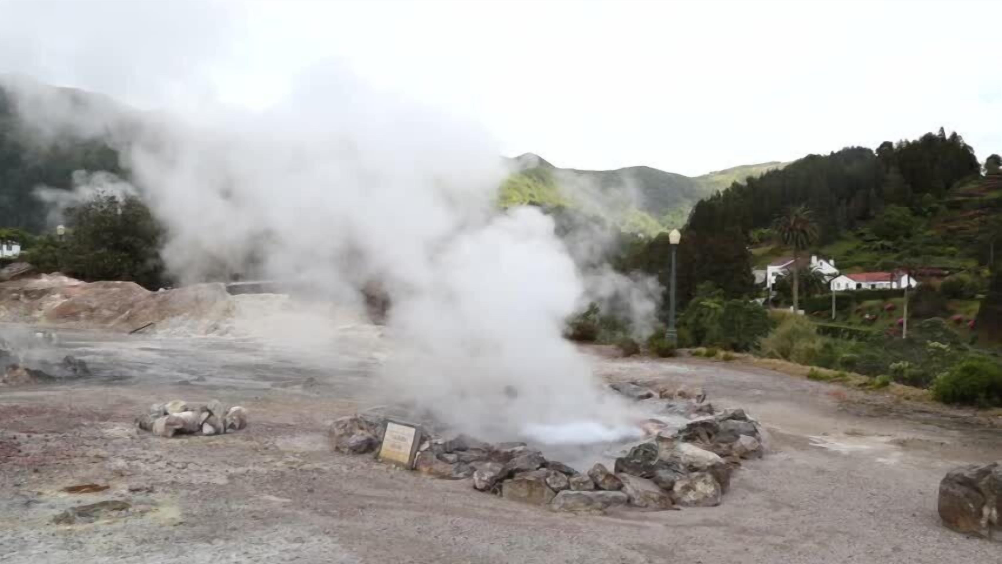 A geothermal area with steam rising from the ground, surrounded by rocky terrain and distant houses.