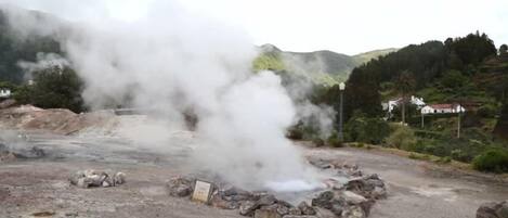 A geothermal area with steam rising from the ground, surrounded by rocky terrain and distant houses.