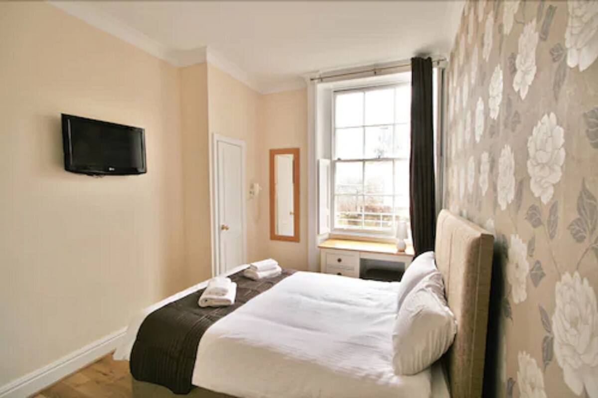 A hotel room with a bed, a television mounted on the wall, a window with curtains, and a floral-patterned wall.