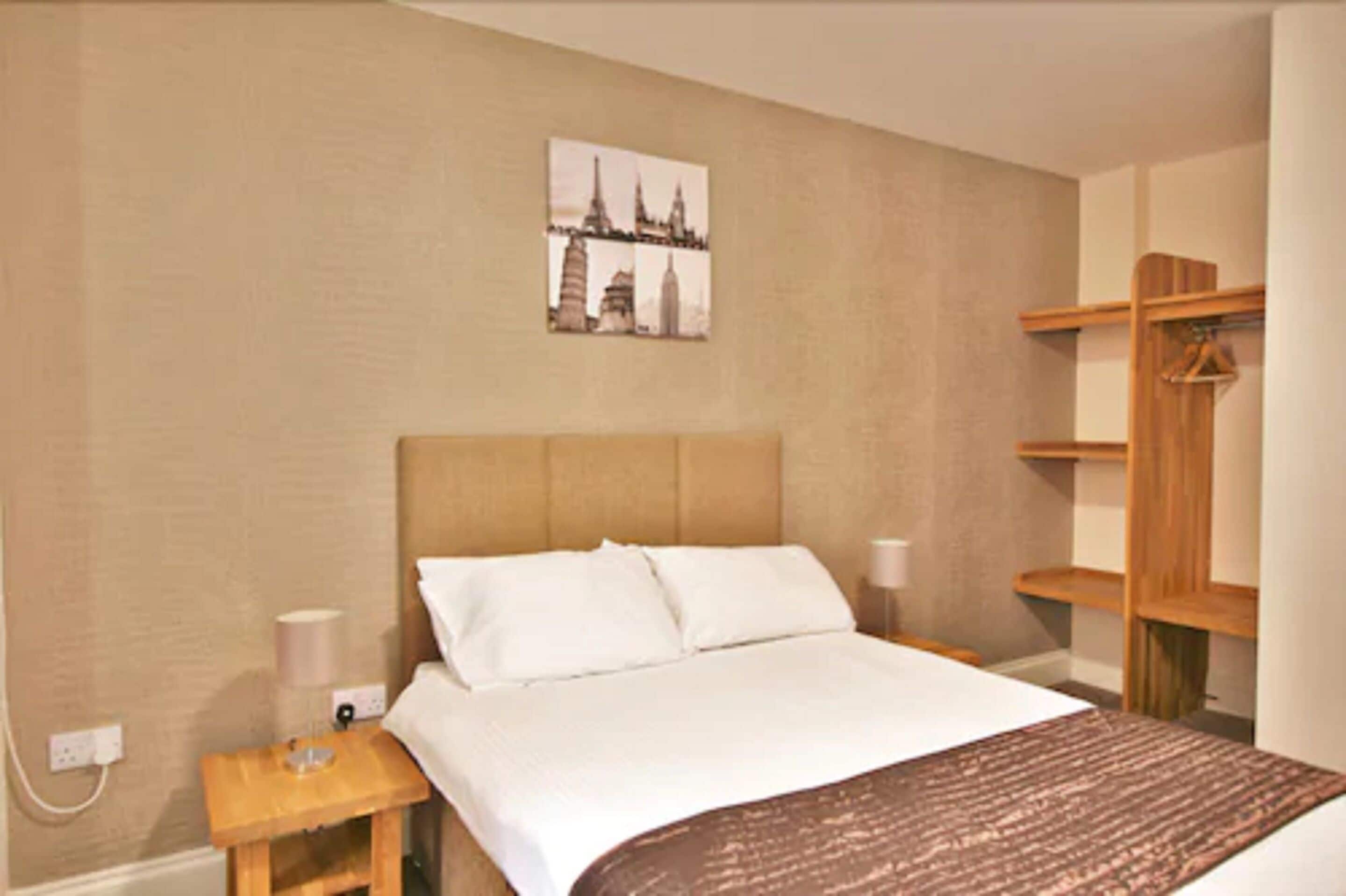 A hotel room with a bed, a nightstand, a lamp, a wooden shelf, and a wall-mounted picture of a cityscape.