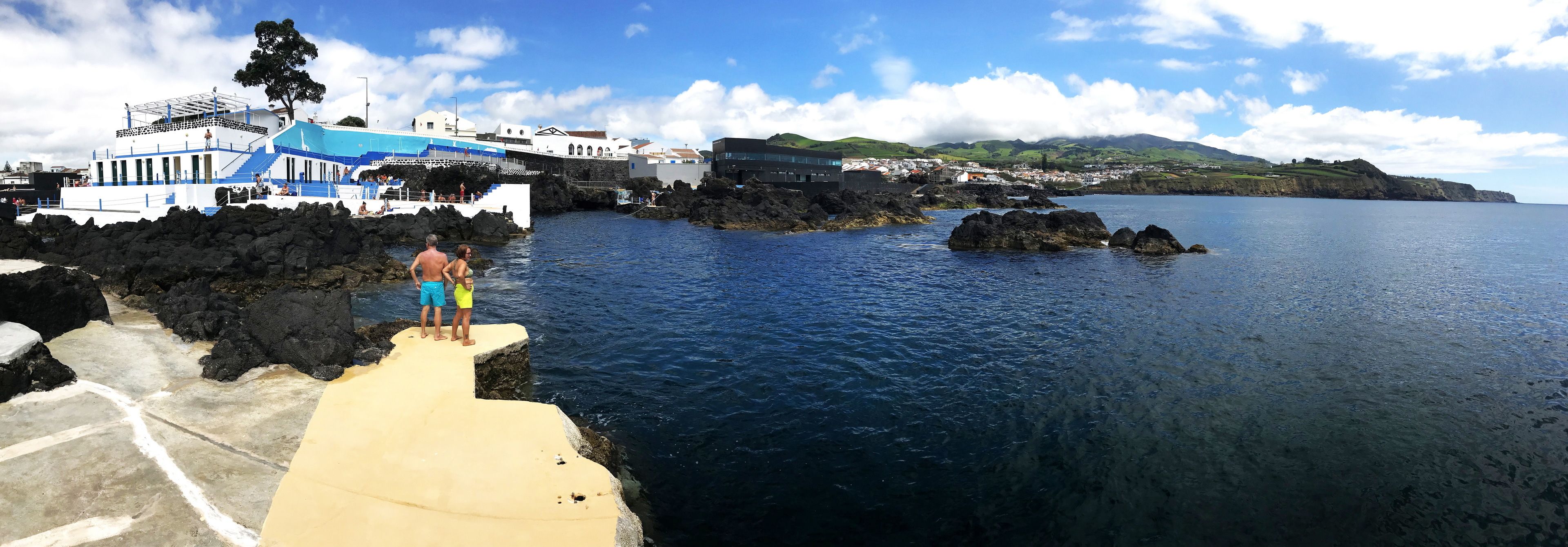 A coastal scene with a pier extending into the water, a building with blue and white architecture, and a town in the background.
