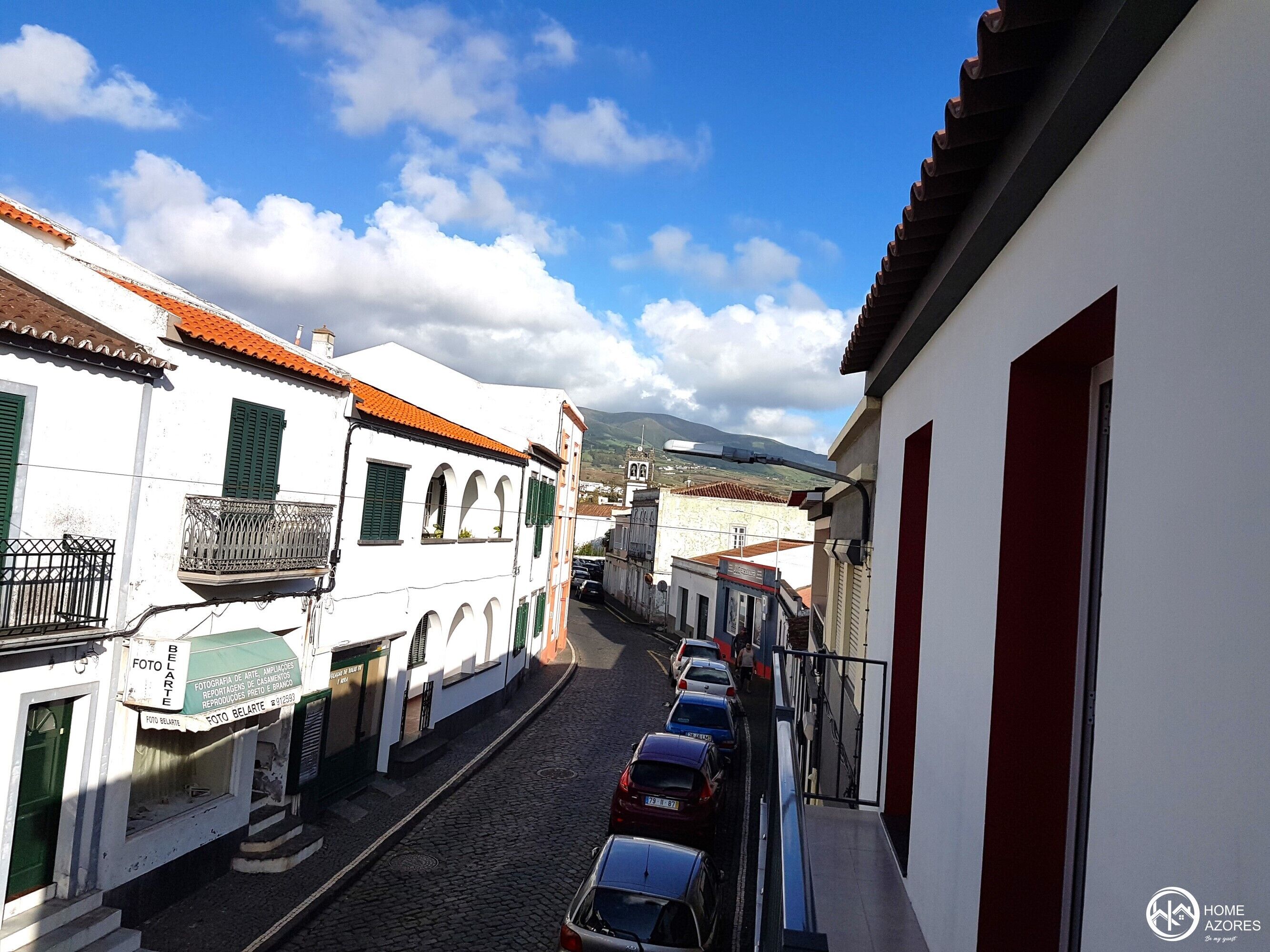Una estrecha calle empedrada bordeada por edificios blancos con contraventanas verdes y techos de tejas rojas.