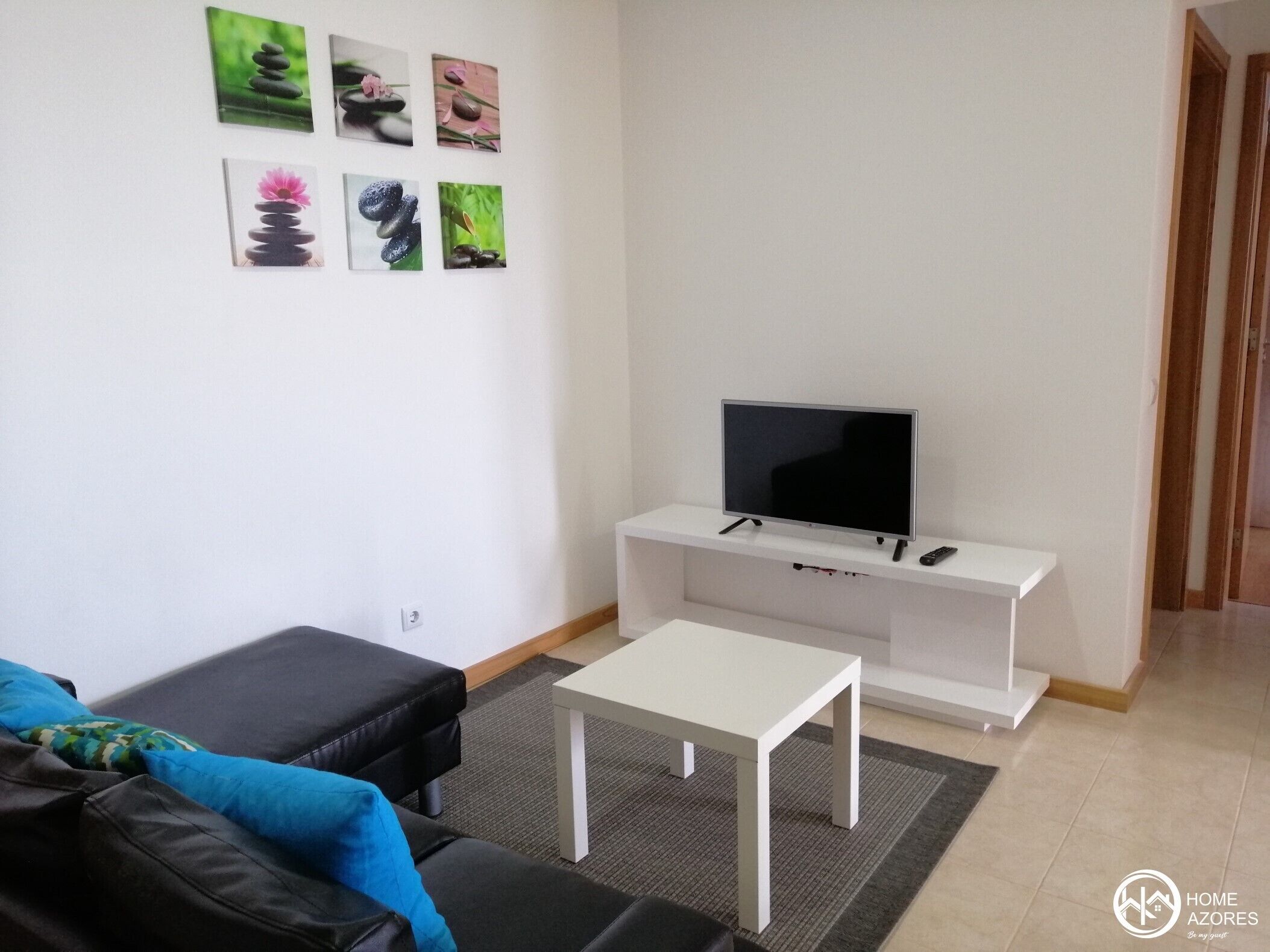 En moderne stue med et fladskærms-tv på et hvidt stativ, en sort lædersofa, et lille hvidt bord og en grå måtte.