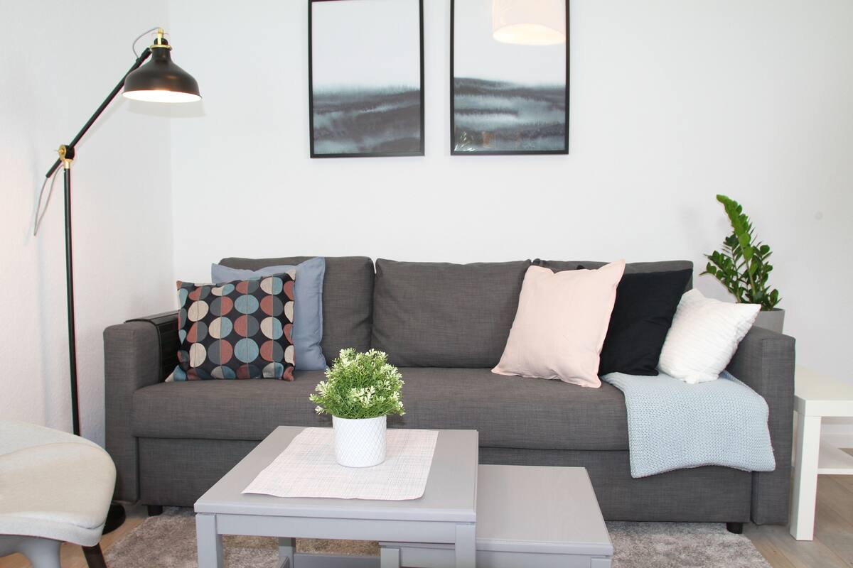 A modern living room with a grey sofa, a small white table, and a floor lamp. There are two framed pictures on the wall above the sofa.