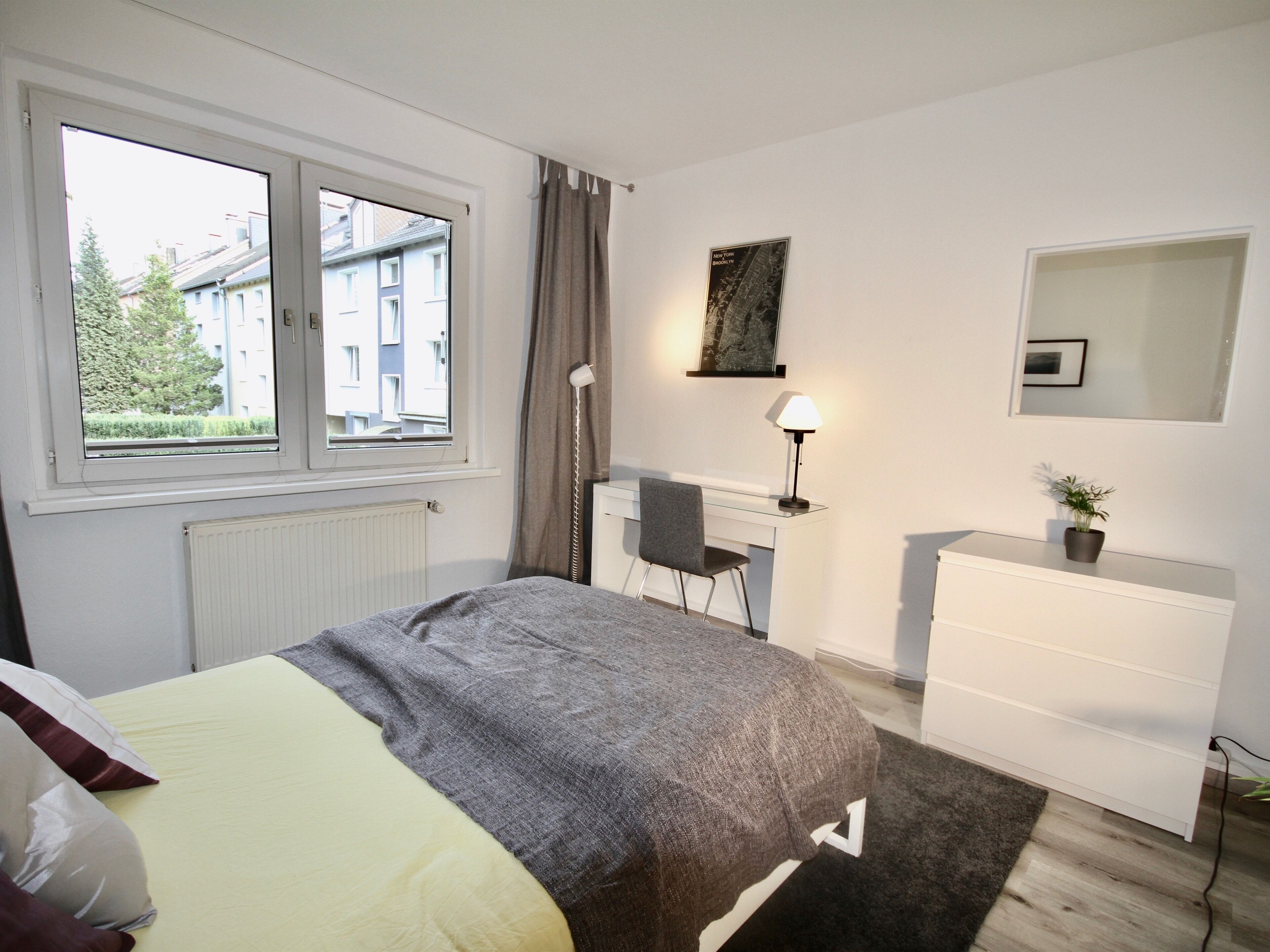 A bedroom with a bed, a desk, a chair, a dresser, a lamp, and a window with a view of buildings.
