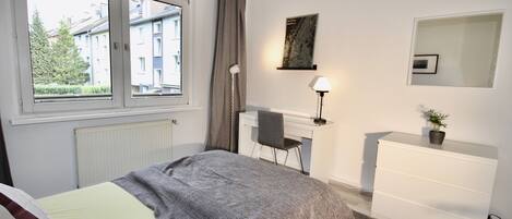 A bedroom with a bed, a desk, a chair, a dresser, a lamp, and a window with a view of buildings.