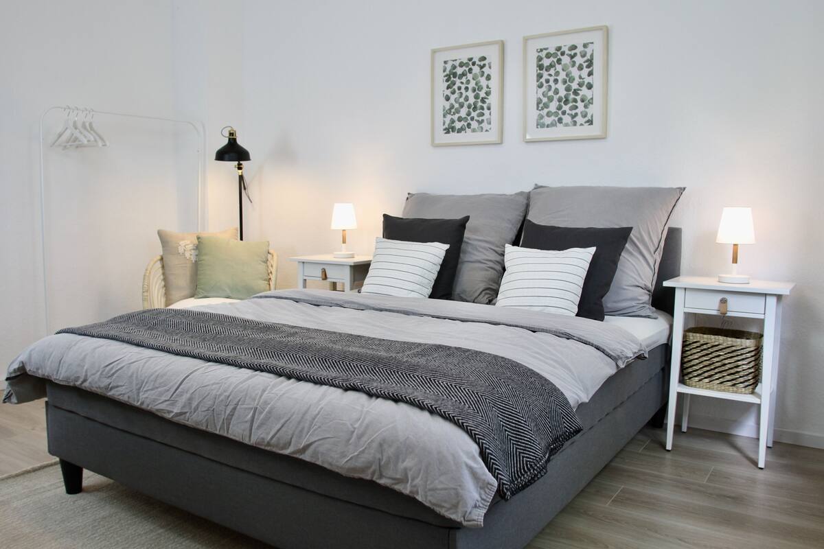 A modern bedroom with a grey bed, two bedside tables with lamps, a white chair, and two framed leaf-patterned wall art pieces.