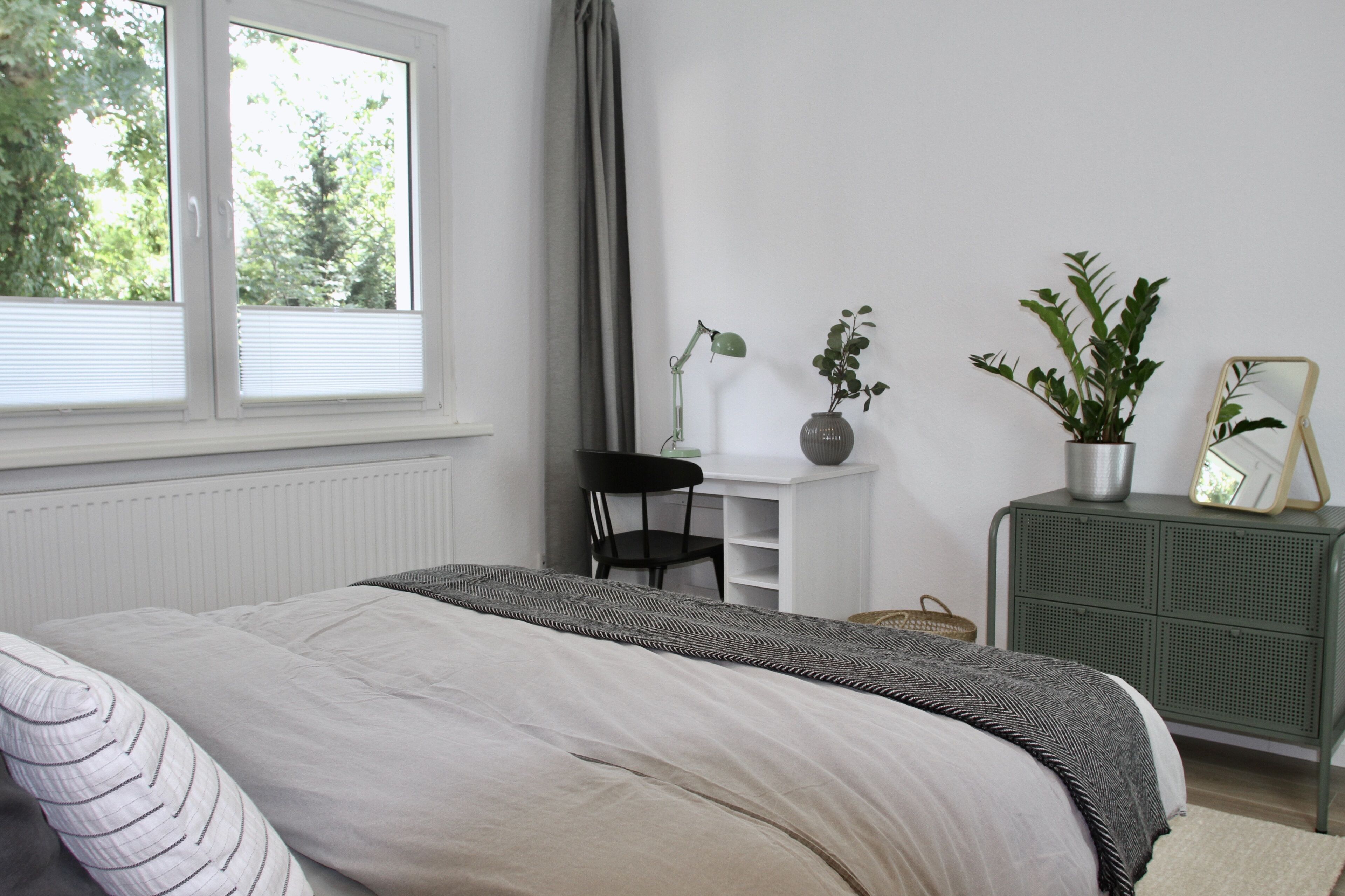 A bedroom with a bed, a desk, a chair, a dresser, and two potted plants.
