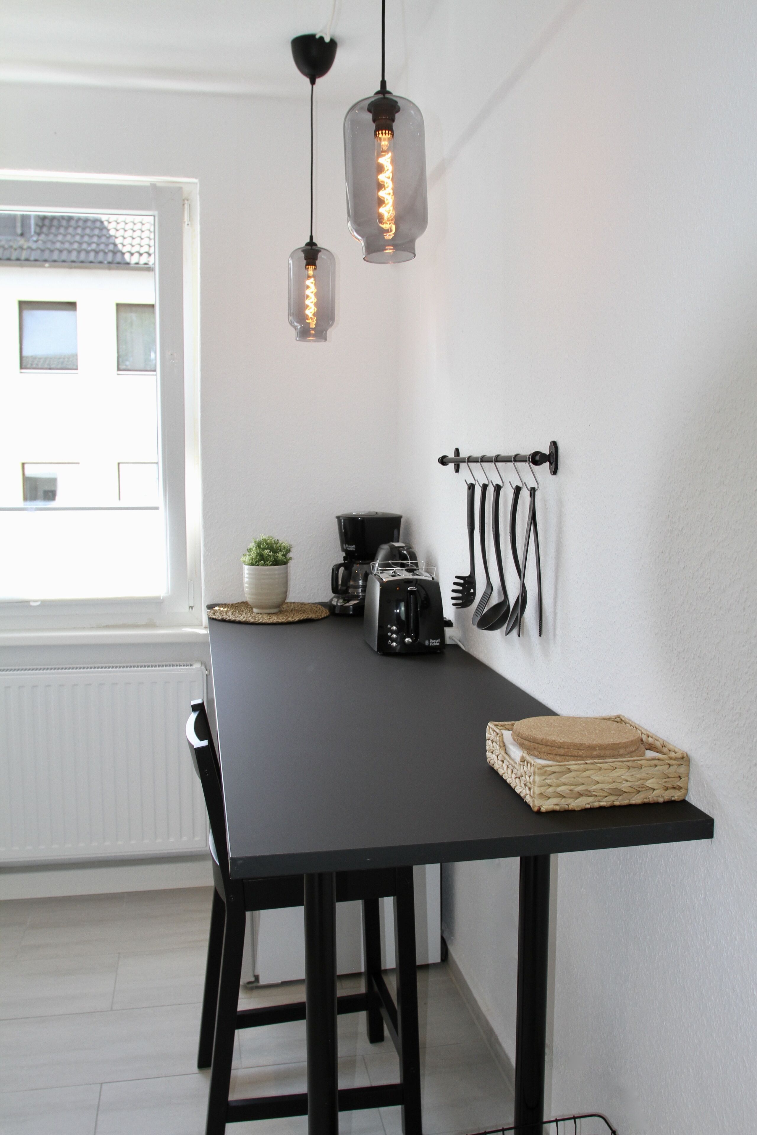 Et køkken med et sort bord, hængende redskaber, en kaffemaskine og en potteplante.