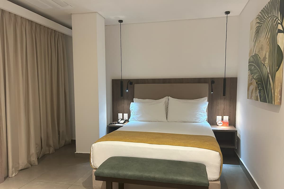 Een hotelkamer met een groot bed, een nachtkastje met een lamp, een bankje en een schilderij aan de muur.