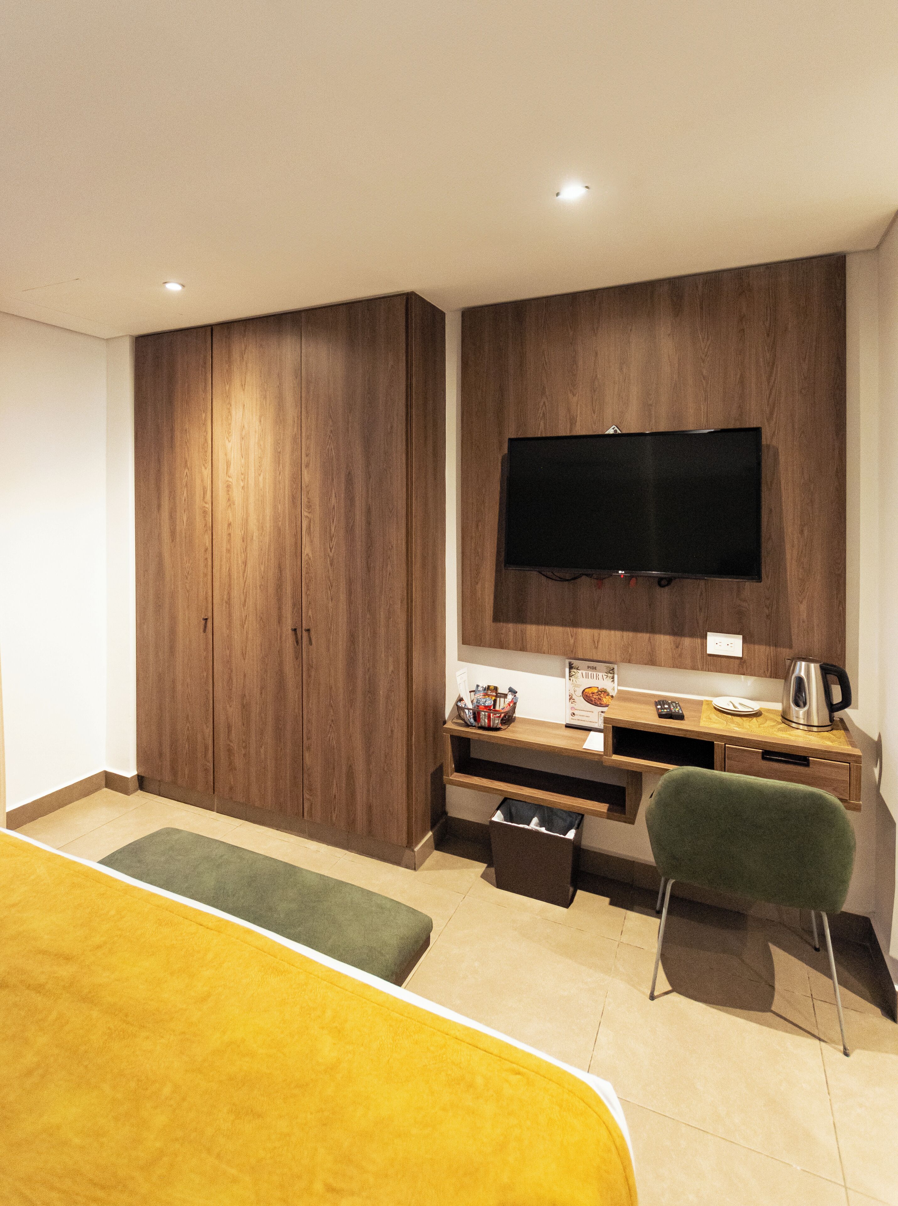 A modern hotel room with a wooden wardrobe, a flat-screen TV mounted on the wall, a desk with a kettle, a green chair, and a yellow bed.