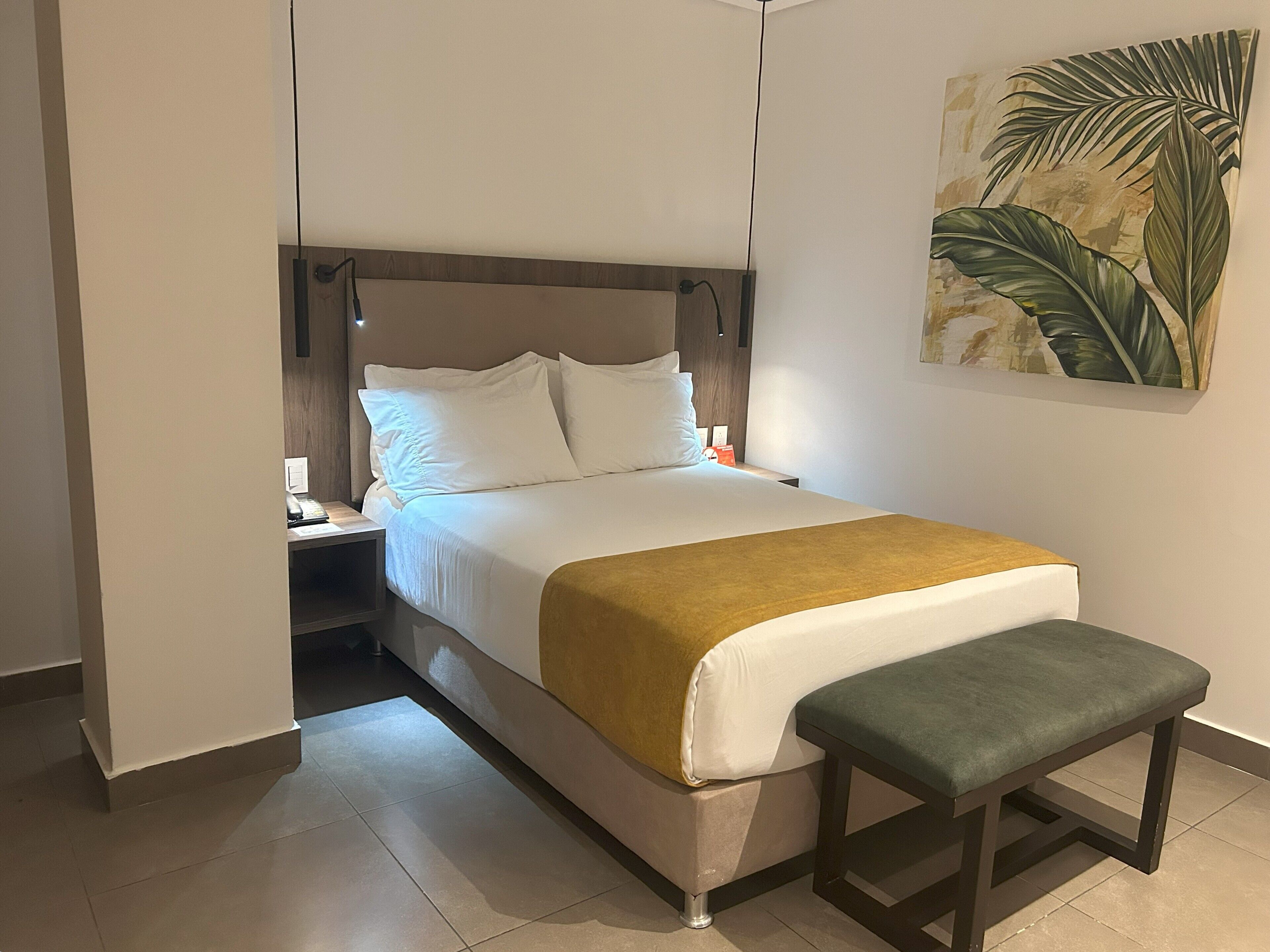 A neatly made bed with white linens and a mustard-colored throw, a wooden bench at the foot of the bed, and a nightstand with a lamp.