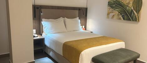 A neatly made bed with white linens and a mustard-colored throw, a wooden bench at the foot of the bed, and a nightstand with a lamp.