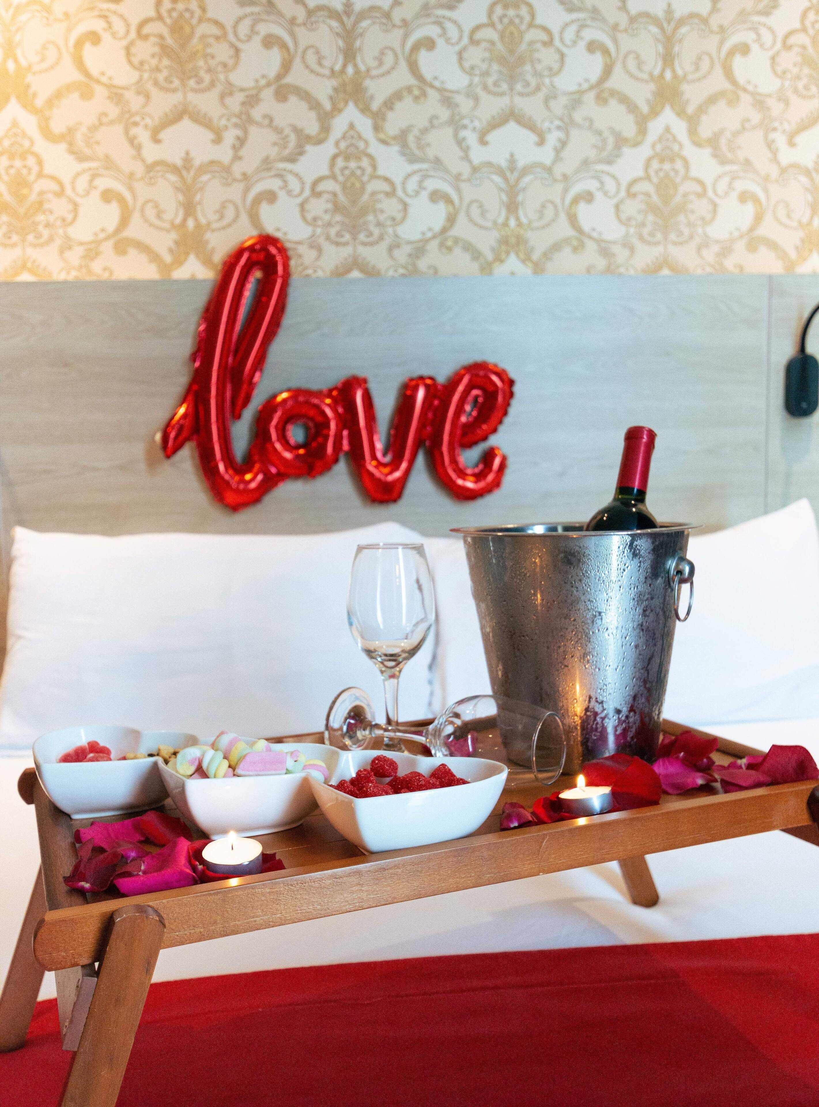 A bed with a decorative wall behind it, a bedside table with a bottle of wine, glasses, and bowls of food.
