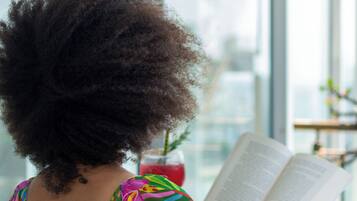 Una persona leyendo un libro en una habitación con ventanas amplias y un sillón estampado.