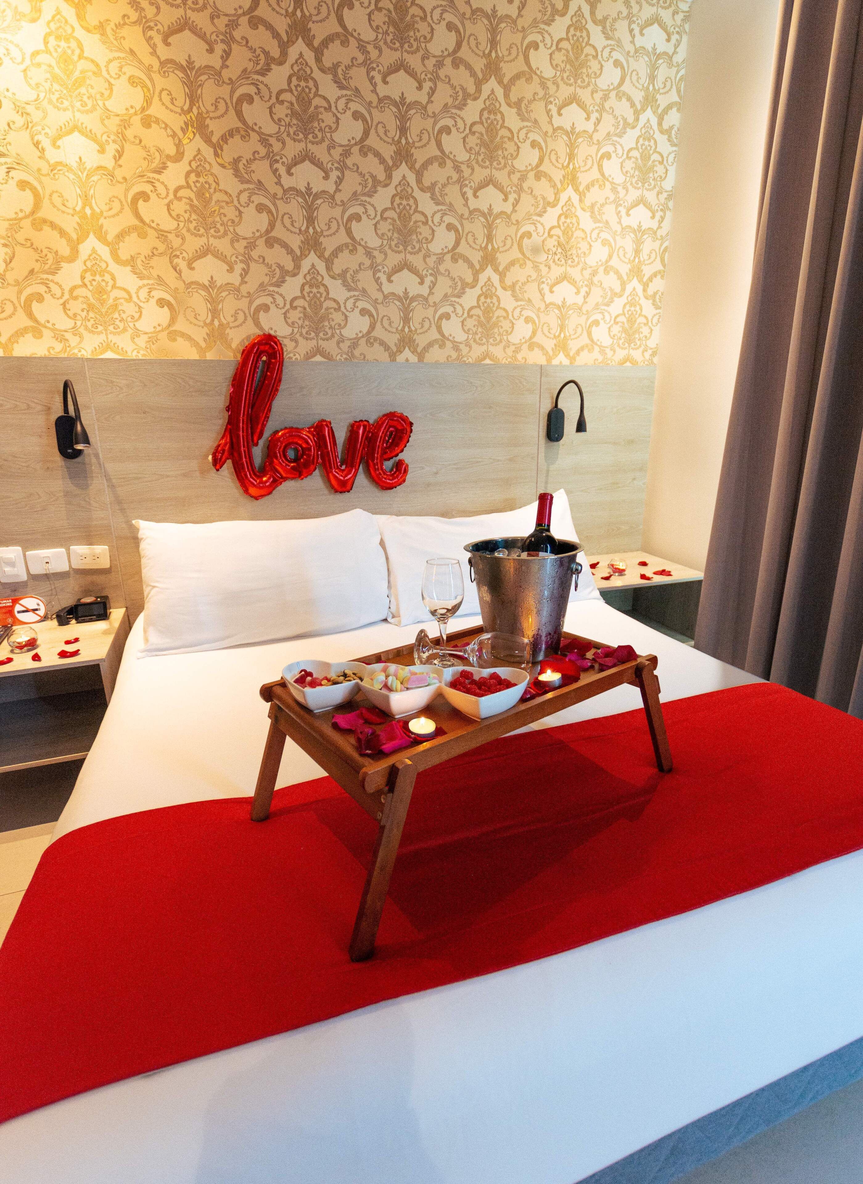 A hotel room with a bed, a bedside table with a lamp, a nightstand, a tray with food and drinks, and a 'love' sign on the headboard.