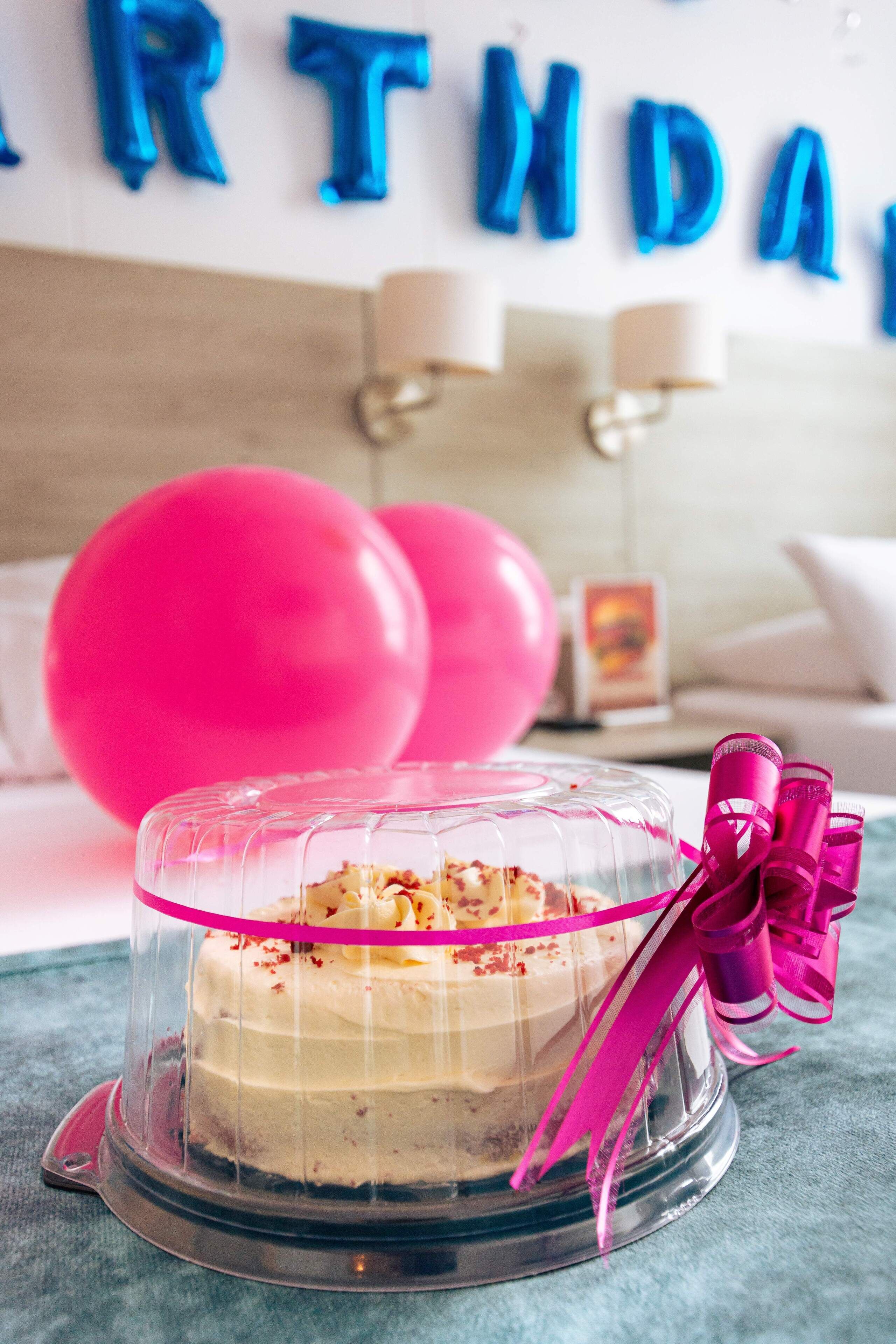 A birthday cake with a clear dome, pink ribbon, and a bow, placed on a table with a blue tablecloth.