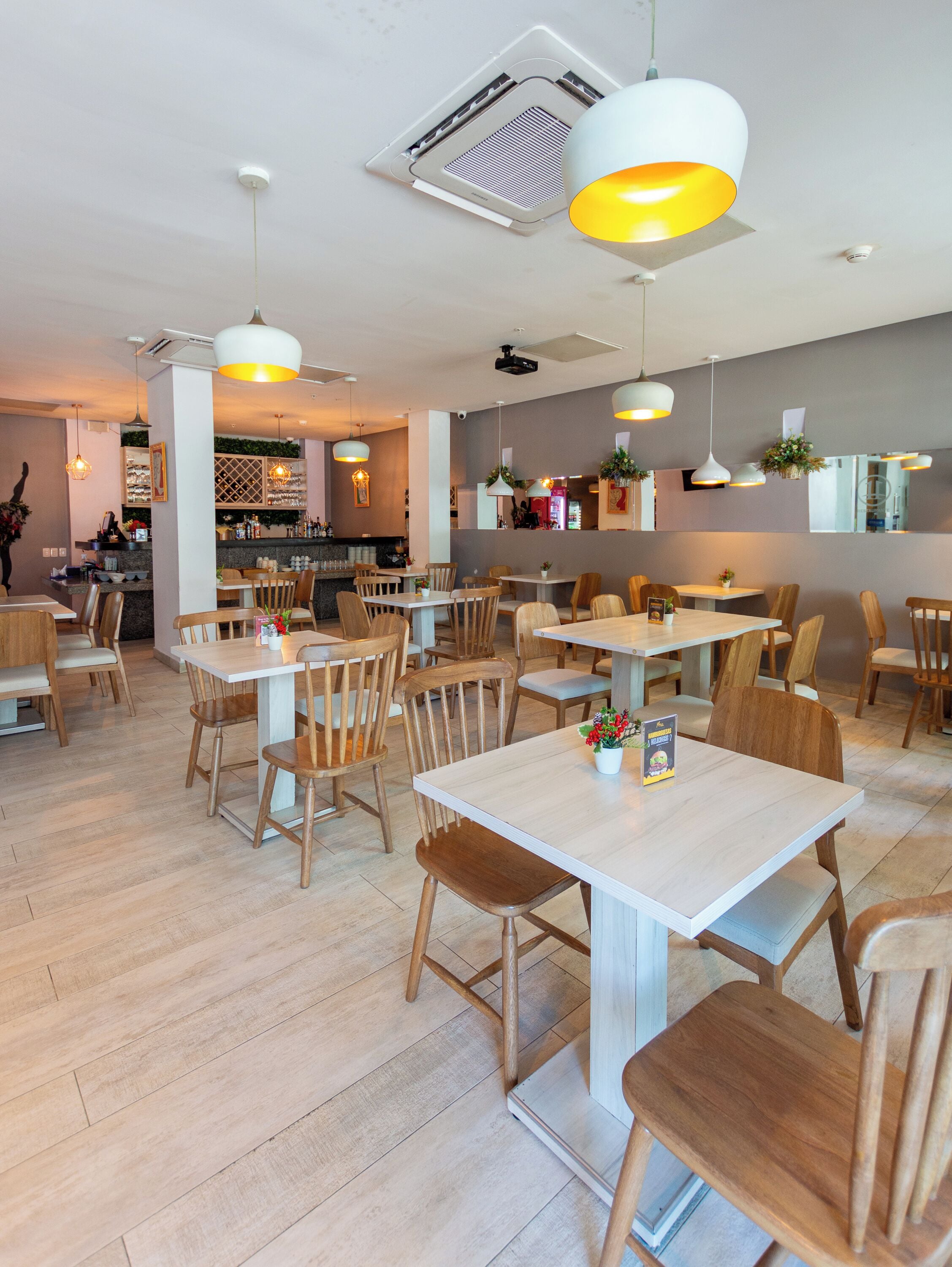 A modern restaurant with wooden tables and chairs, pendant lighting, and a visible kitchen area.