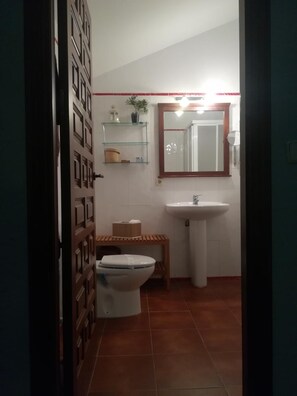 A bathroom with a toilet, sink, and mirror. There are shelves above the sink and a bench below the toilet. - Hostal la Cerca (Arcones)