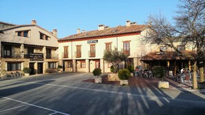 A street view with a building labeled "HOSTAL" featuring a central entrance, a small outdoor seating area, and a parking lot. - Hostal la Cerca (Arcones)
