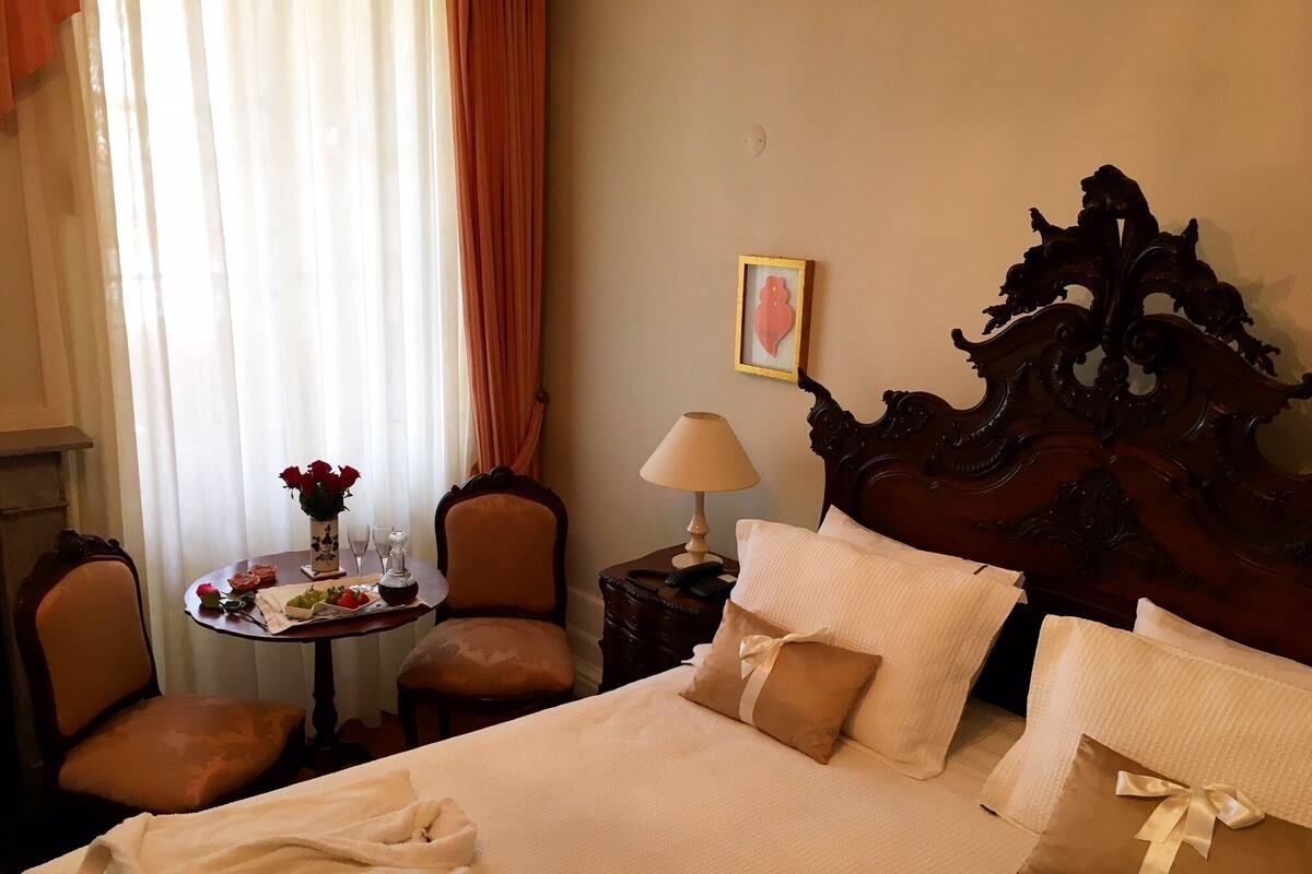 Una habitación de hotel con una cama grande, una mesita de noche con lámpara, una silla, una pequeña mesa con flores y una ventana con cortinas.