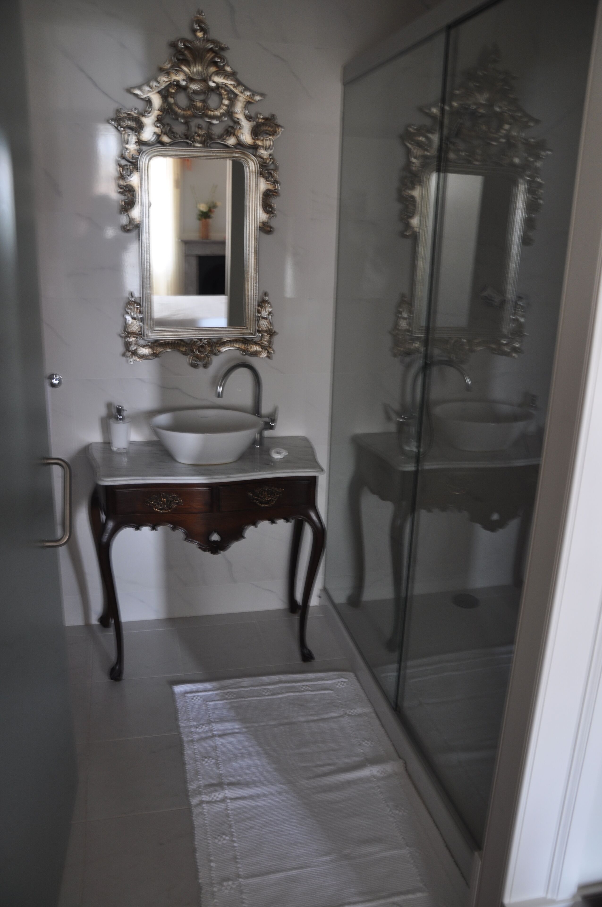 Une salle de bain avec un mur en marbre, un lavabo blanc, un miroir doté d’un cadre orné et une cabine de douche en verre.