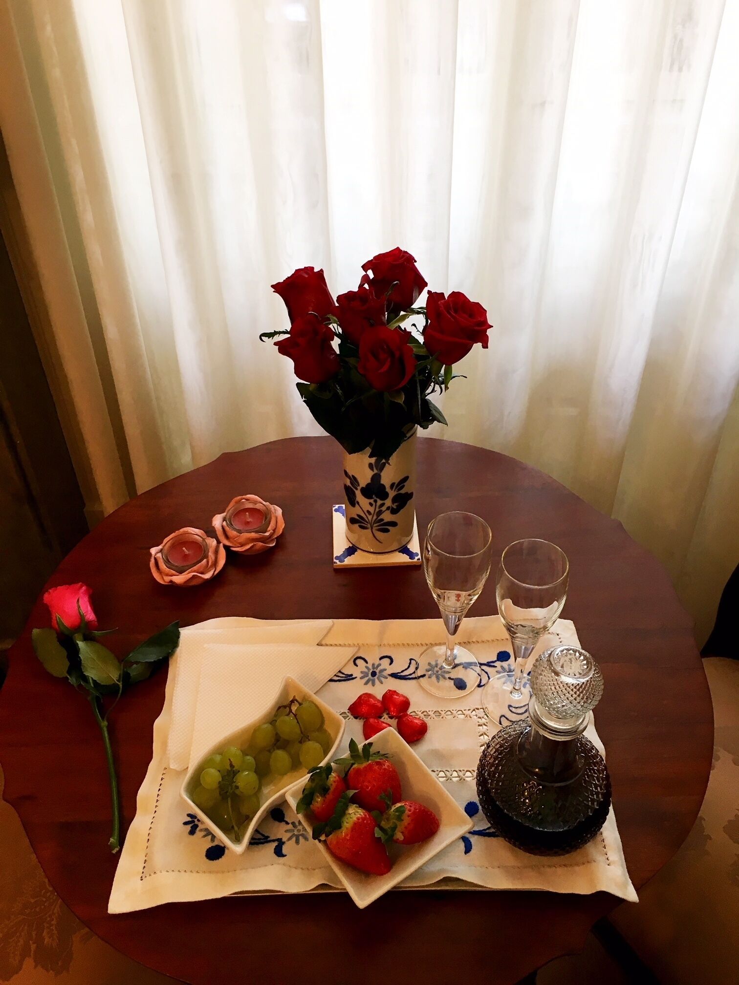 Une table à manger ornée d’un vase de roses rouges, de deux bougies, d’un verre d’eau, d’un verre de vin, d’une assiette de raisins et de fraises, ainsi que d’un salier et d’un poivrier.