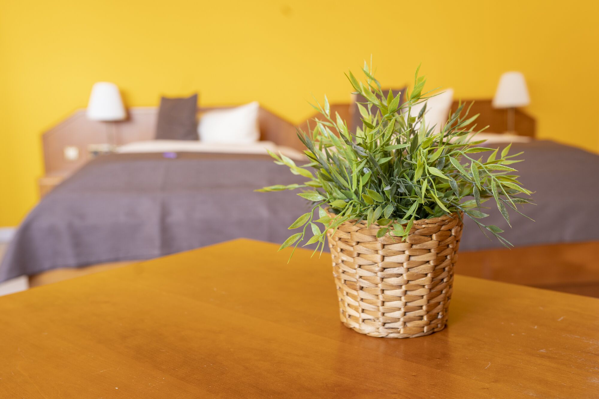 A bed with a grey bedspread and white pillows, a bedside table with a woven plant pot, and a yellow wall in the background.