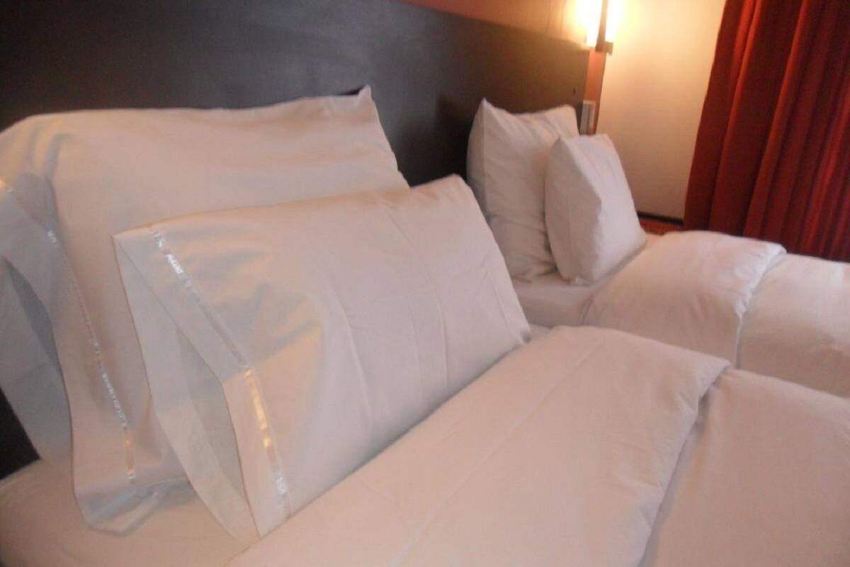 Una cama bien hecha con sábanas y almohadas blancas, un cabecero y una lámpara fijada a la pared.