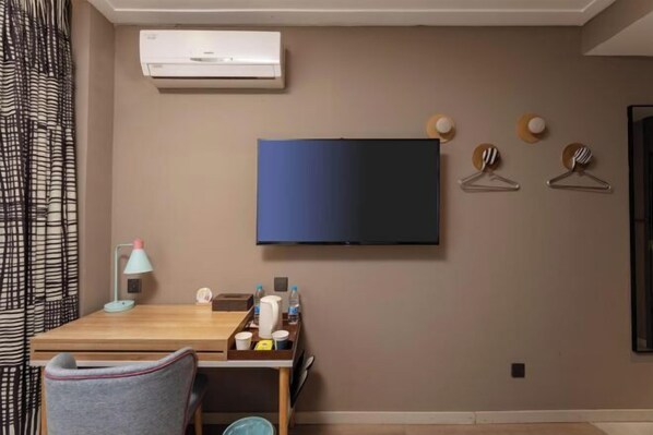Una habitación con una televisión de pantalla plana montada en la pared, un escritorio con lámpara, una silla y una mesita con objetos personales.