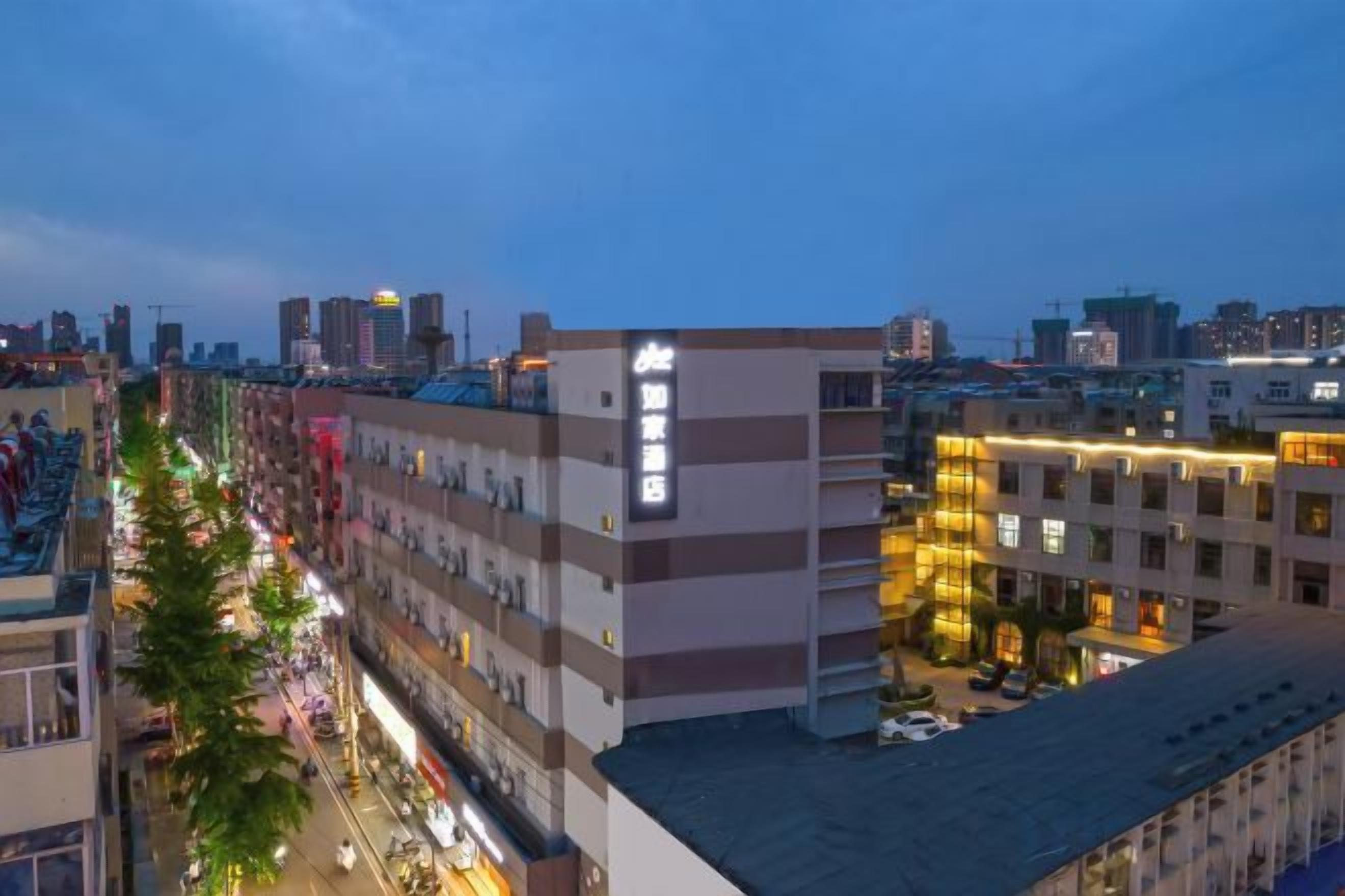 A multi-story hotel with a row of rooms on the ground floor, a parking area, and a view of the city skyline.