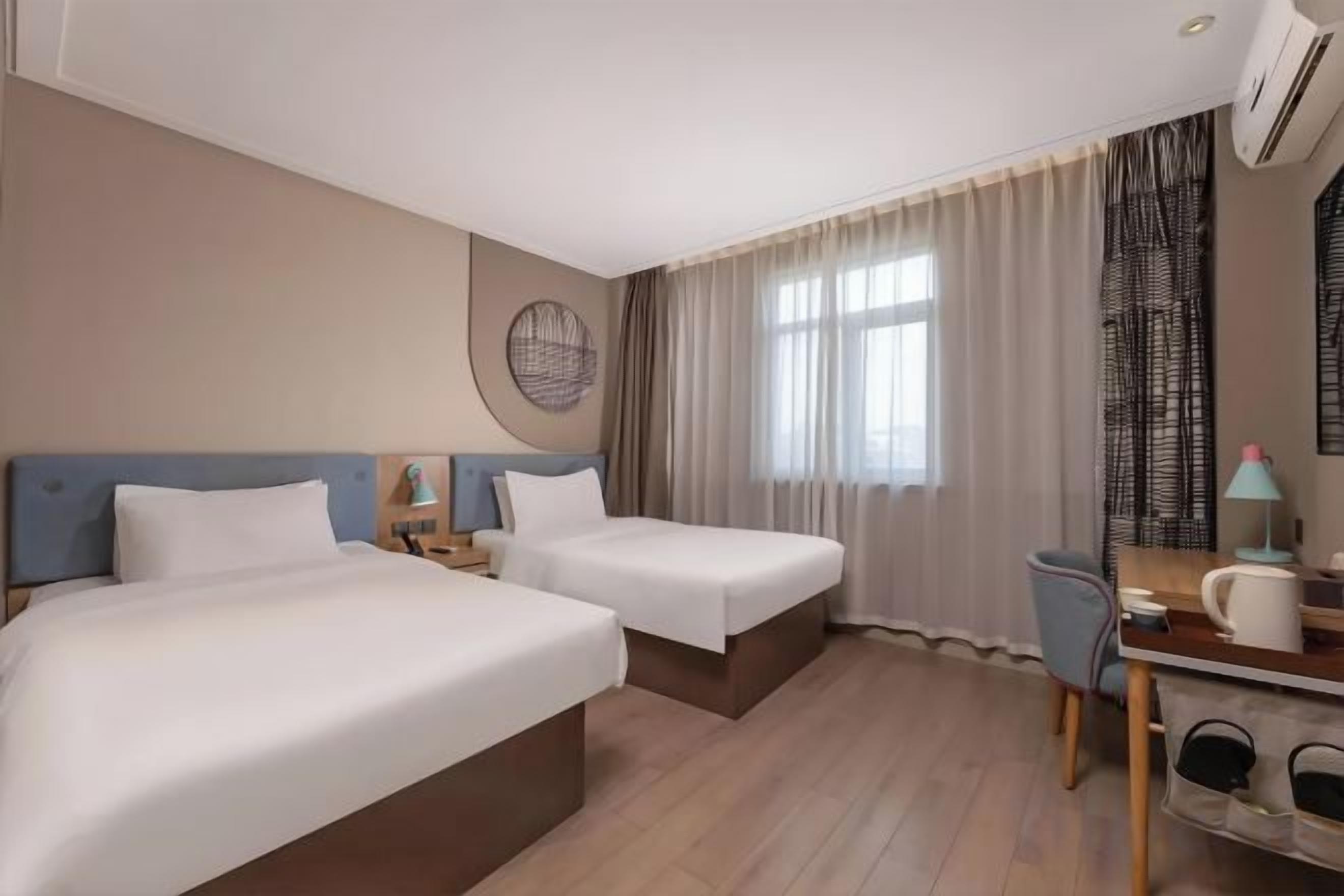 A hotel room with two beds, a desk, a chair, a window with curtains, and a wall-mounted air conditioner.