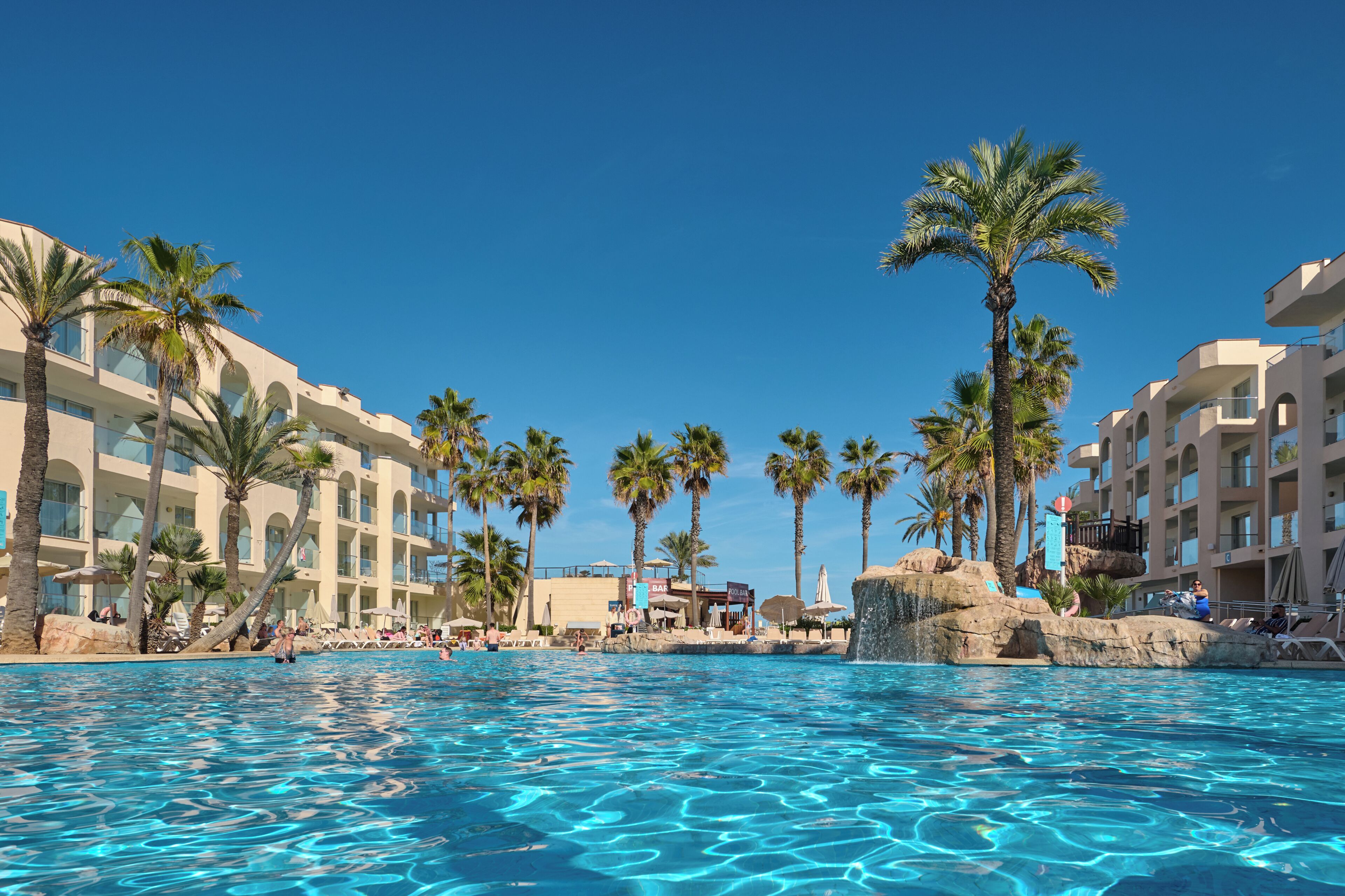 A swimming pool with clear water, surrounded by palm trees and a resort building with multiple balconies.