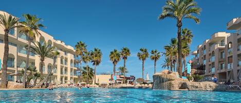 A swimming pool with clear water, surrounded by palm trees and a resort building with multiple balconies.