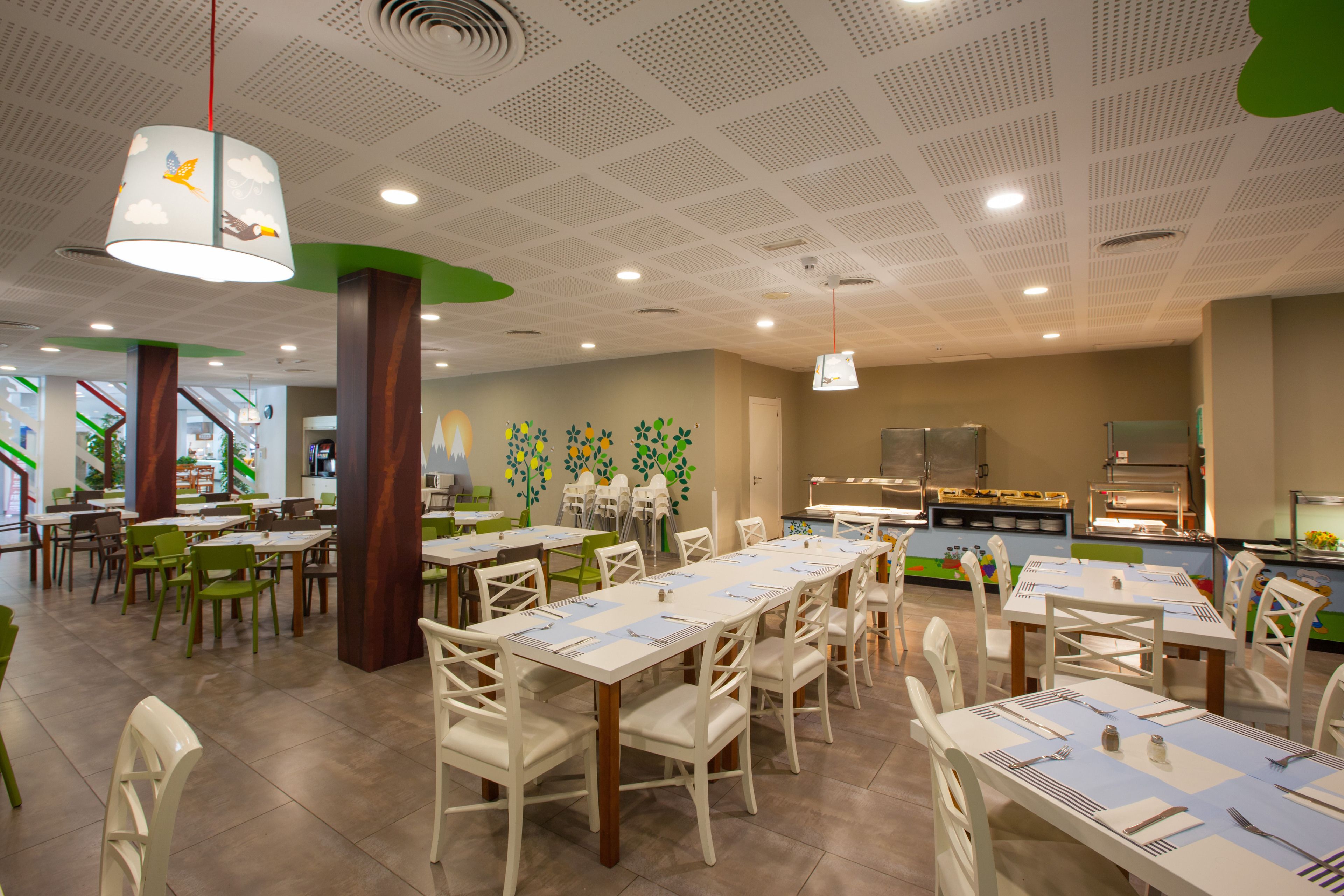 Un comedor con mesas dispuestas para el servicio, un área de juegos para niños al fondo y una cocina visible a través de una puerta abierta.