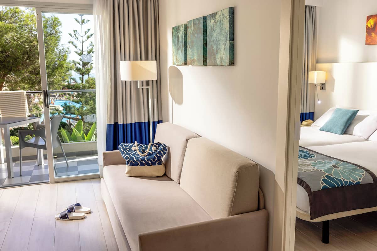 A hotel room with a sofa, a bed, a balcony with a view of greenery and a pool, and a table with a chair.