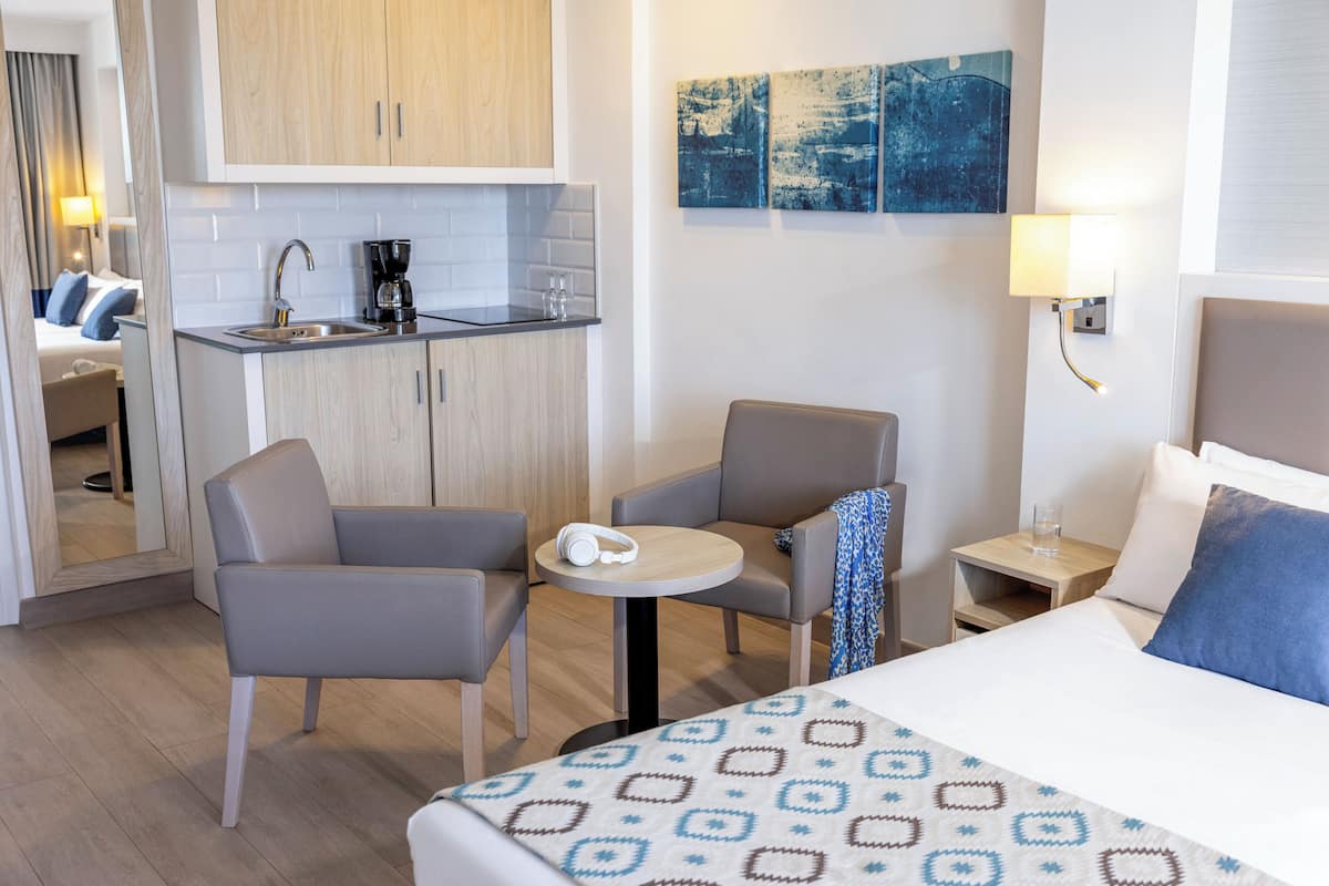 A hotel room with a bed, two armchairs, a small table, a kitchenette, and a nightstand with a lamp.