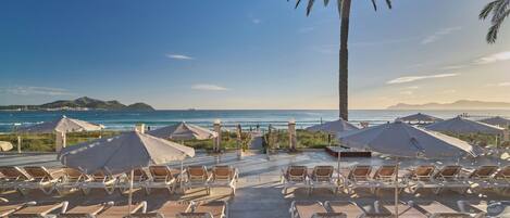 A beachside resort with sun loungers and umbrellas, a palm tree, and a view of the ocean and distant mountains.
