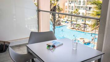 Un balcón con una mesa, un par de gafas de sol y un vaso de agua. Se ve una piscina con gente y palmeras al fondo.