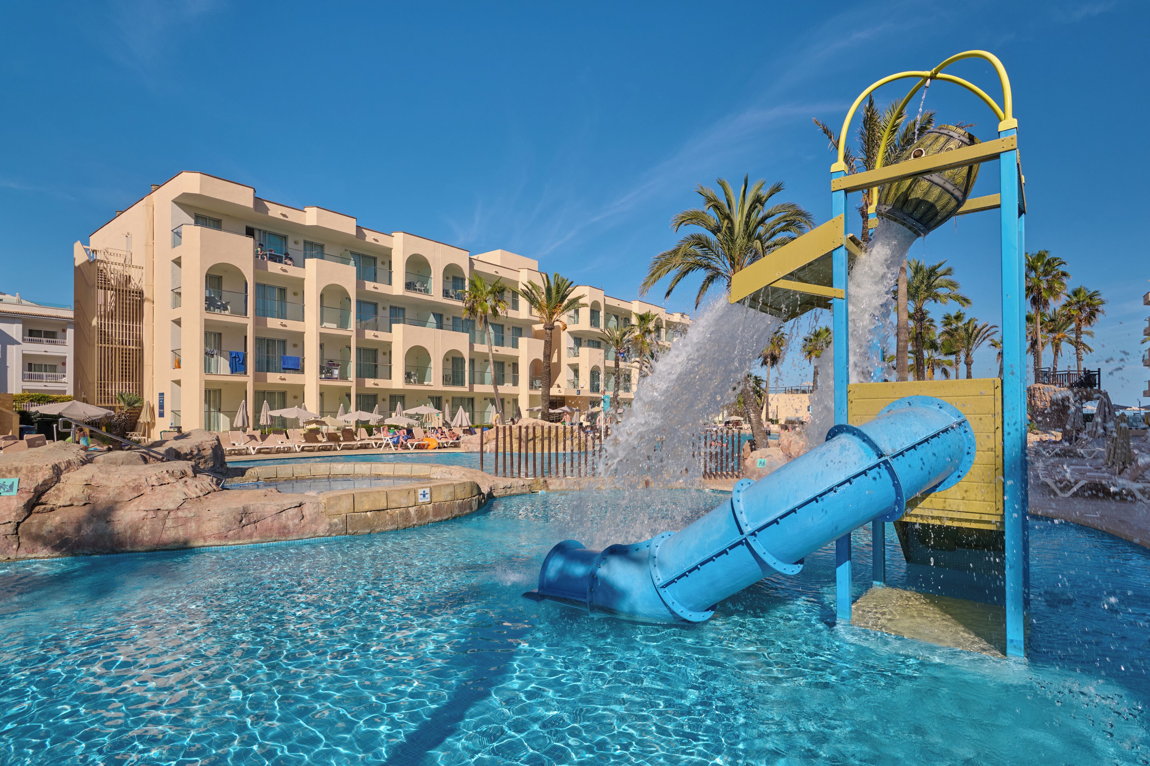 A hotel pool with a water slide, palm trees, and a clear blue sky.