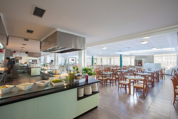 A spacious dining area with multiple tables and chairs, a buffet counter with various food items, and a modern kitchen in the background.